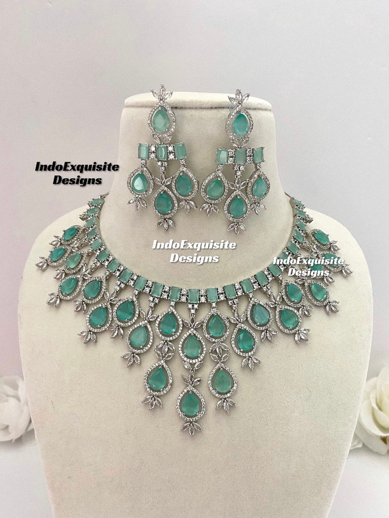 May include: A silver necklace and earrings set with teardrop-shaped light green stones and clear rhinestones. The necklace has a V-shaped design and the earrings are dangle style. The set is labeled "IndoExquisite Designs".