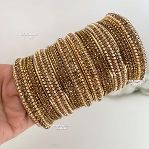 Peut inclure: Une pile de bracelets dorés avec des détails complexes. Les bracelets sont ornés de petites perles rondes blanc cassé et de cristaux bruns clairs scintillants. Le motif est répété sur plusieurs bracelets, créant un effet superposé.