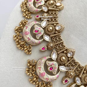 Pu&ograve; includere: Primo piano di una collana dorata con dettagli intricati. La collana presenta pendenti a forma di mezzaluna con disegni floreali rosa e bianchi, accentuati da pietre rosse. Il design include perline dorate pendenti e pietre chiare a forma di goccia.