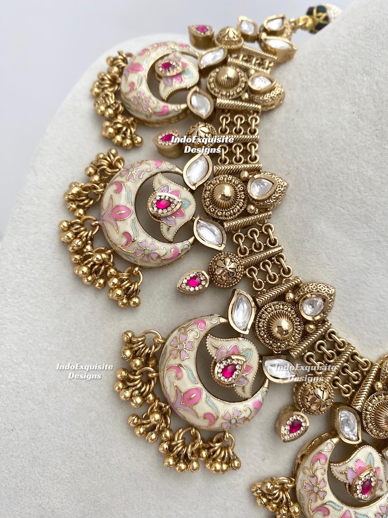 Pu&ograve; includere: Primo piano di una collana dorata con dettagli intricati. La collana presenta pendenti a forma di mezzaluna con disegni floreali rosa e bianchi, accentuati da pietre rosse. Il design include perline dorate pendenti e pietre chiare a forma di goccia.