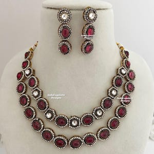 Peut inclure: Un ensemble de collier à deux rangs et de boucles d'oreilles assorties. Les bijoux présentent des pierres ovales de couleur rubis entourées de petits cristaux transparents. Le métal est de couleur dorée. Les boucles d'oreilles ont une pierre ronde supérieure et trois pierres couleur rubis.