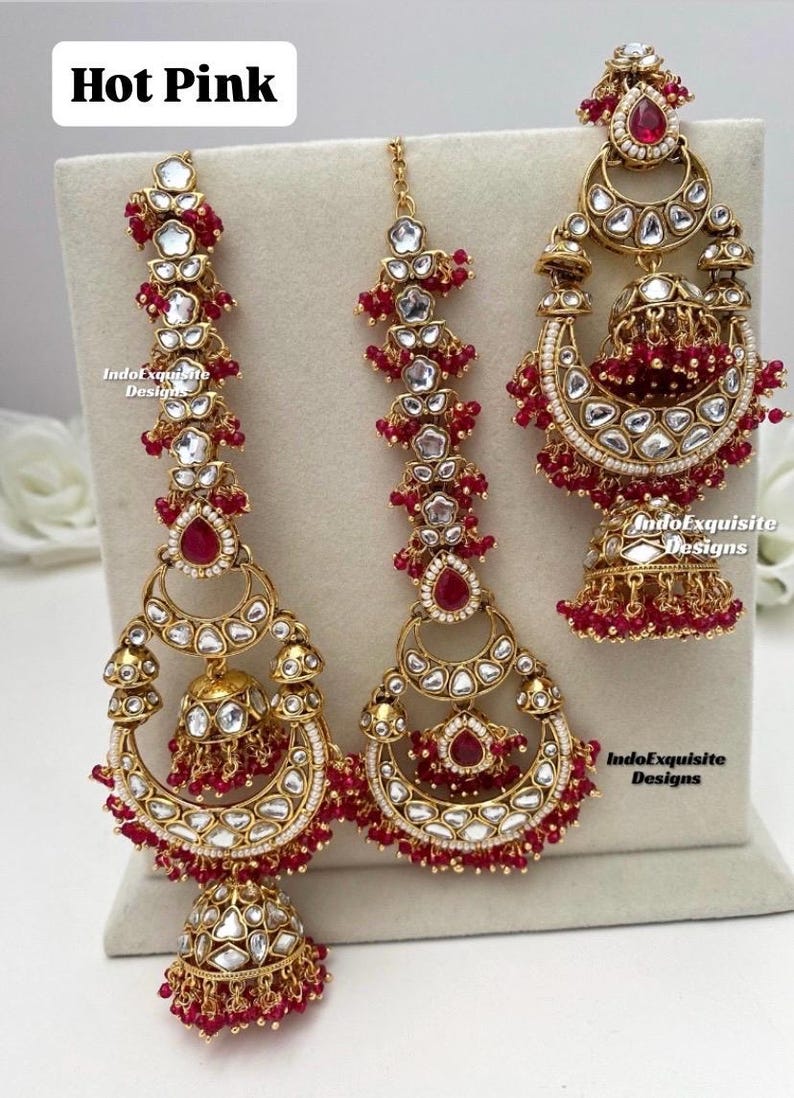 Puede incluir: Un conjunto de joyer&iacute;a etiquetado como Hot Pink que incluye dos grandes pendientes dorados y un adorno para la cabeza a juego. Cada pieza est&aacute; decorada con piedras claras, piedras rojas en forma de l&aacute;grima y peque&ntilde;as cuentas rojas, con formas de media luna y colgantes jhumka.