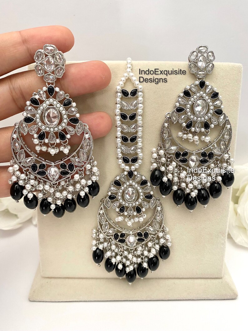 May include: A set of silver earrings and a matching maang tikka, featuring black and white stones and pearls. The earrings have a crescent moon shape with dangling black beads. The maang tikka has a similar design with a long chain of pearls.