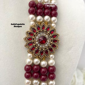 May include: A multi-strand necklace featuring alternating rows of white faux pearls and burgundy beads. The focal point is a gold-toned floral pendant with red gemstones and a central clear stone. The necklace is displayed on a white mannequin.