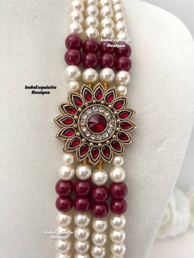 May include: A multi-strand necklace featuring alternating rows of white faux pearls and burgundy beads. The focal point is a gold-toned floral pendant with red gemstones and a central clear stone. The necklace is displayed on a white mannequin.