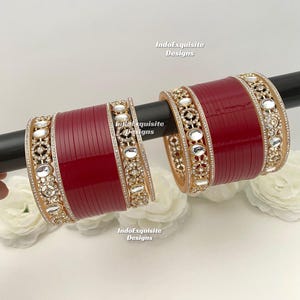 May include: Two maroon bangles with gold and crystal embellishments. The bangles are displayed on a black rod, with white flowers in the background. The bangles have a repeating pattern of crystals and gold filigree.