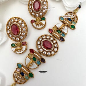 May include: Gold-toned jewelry set featuring oval-shaped ruby-colored stones surrounded by clear crystals. Additional pieces include pendants with a tiger design on a white background, and colorful gemstones. The set is displayed on a white surface.