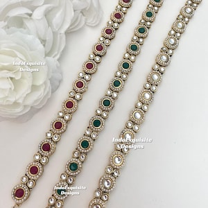 Może przedstawiać: Trzy złote łańcuszki z białymi perełkami i kolorowymi kamieniami szlachetnymi. Łańcuszki mają czerwone, zielone i przezroczyste kamienie szlachetne. Łańcuszki leżą na białym tle.