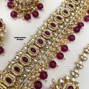 May include: A gold necklace with a large, ornate pendant featuring a central ruby-red stone surrounded by smaller white stones. The necklace is adorned with dangling ruby-red beads and white pearls. The pendant is surrounded by a delicate gold filigree design. The necklace is a traditional Indian style.