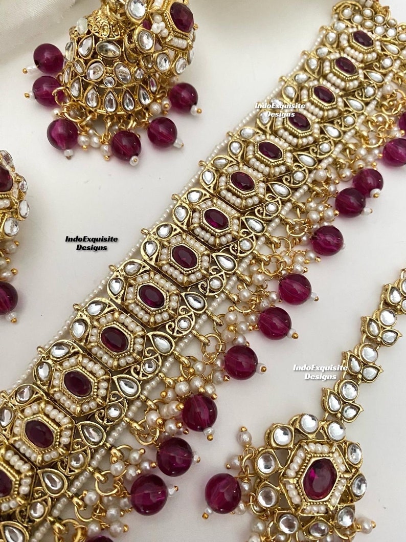 May include: A gold necklace with a large, ornate pendant featuring a central ruby-red stone surrounded by smaller white stones. The necklace is adorned with dangling ruby-red beads and white pearls. The pendant is surrounded by a delicate gold filigree design. The necklace is a traditional Indian style.