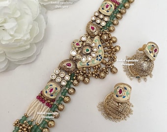 Puede incluir: Un conjunto de collar y pendientes jhumka en tono dorado con cuentas blancas, cuentas verdes y detalles en rosa. La joyería incluye metalistería intrincada, detalles en esmalte y pequeños dijes dorados colgantes. El texto dice IndoExquisite Designs.