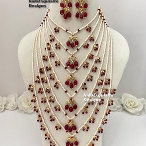 Può includere: Una collana multifilo con perline bianche e accenti di pietre preziose rosse. La collana presenta un design a cascata con più strati di perline e pietre preziose rosse pendenti. La collana è abbinata a orecchini abbinati con un design simile.