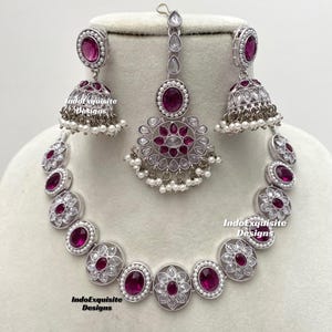 Op de afbeelding: Een zilveren ketting met een roze stenen hanger en bijpassende oorbellen en een maang tikka. De ketting heeft een bloemmotief met witte stenen.