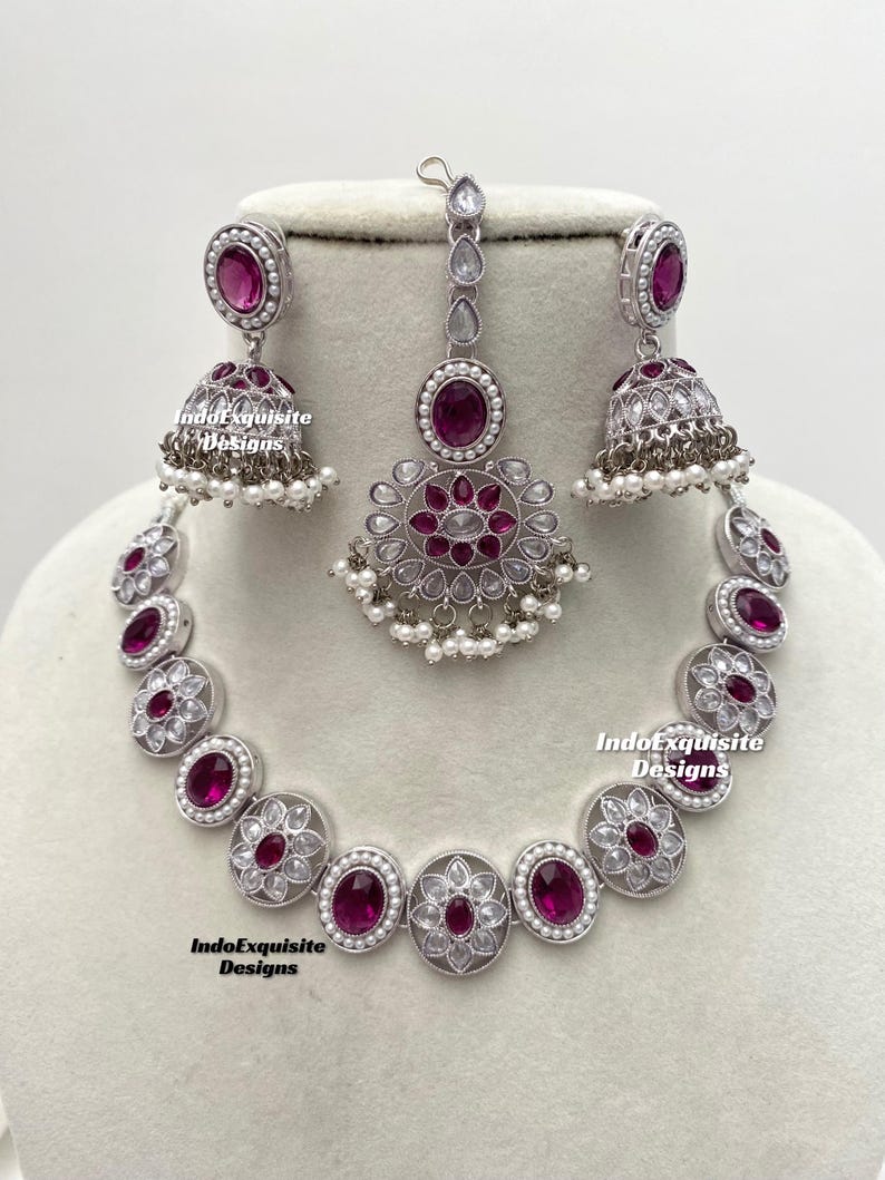 Op de afbeelding: Een zilveren ketting met een roze stenen hanger en bijpassende oorbellen en een maang tikka. De ketting heeft een bloemmotief met witte stenen.