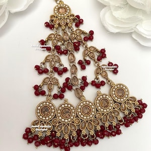 May include: A detailed, gold-coloured headpiece with a complex design. It incorporates circular and teardrop shapes, embellished with small, red beads and clear, round accents. The design includes a central, circular feature and hanging strands.