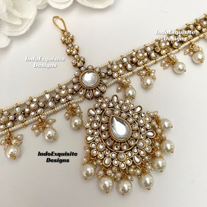 May include: A gold-tone bridal headpiece with a large teardrop-shaped crystal and pearl accents. The headpiece is decorated with small, round crystals and has a delicate chain.