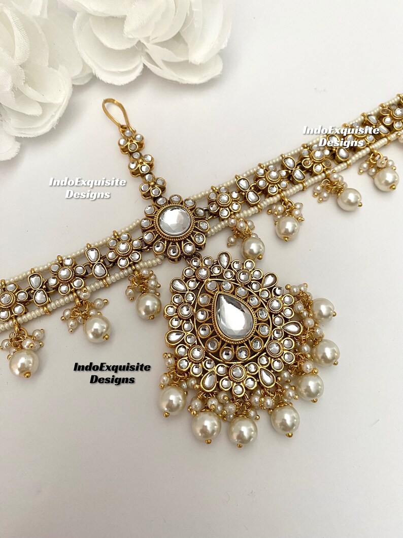May include: A gold-tone bridal headpiece with a large teardrop-shaped crystal and pearl accents. The headpiece is decorated with small, round crystals and has a delicate chain.