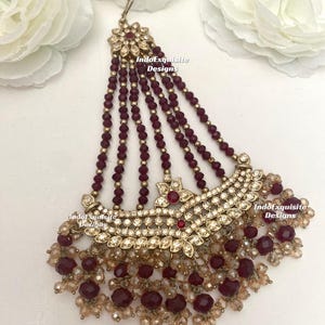 Op de afbeelding: Een sierlijk, goudkleurig hoofddeksel met bordeauxrode en heldere kristallen kralen. Het ontwerp heeft een bloemmotief met sprankelende strass-steentjes en bungelende kralenstrengen. De tekst "IndoExquisite Designs" is zichtbaar.
