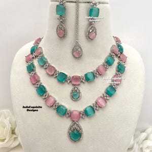 May include: A silver necklace set with a double strand of alternating pink and teal square stones. The necklace has a teardrop pendant with a teal stone. Matching earrings with teal square stones and teardrop pendants are included. The set is labeled "IndoExquisite Designs".