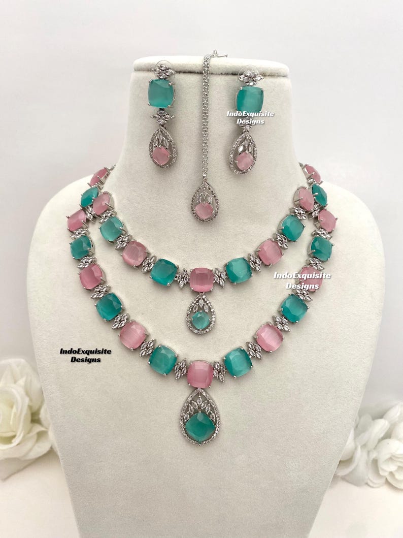 May include: A silver necklace set with a double strand of alternating pink and teal square stones. The necklace has a teardrop pendant with a teal stone. Matching earrings with teal square stones and teardrop pendants are included. The set is labeled "IndoExquisite Designs".