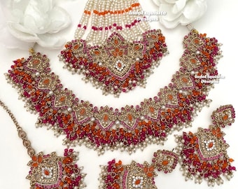 May include: A traditional Indian bridal jewelry set featuring a gold-toned necklace, earrings, and a headpiece. The set is adorned with pink, orange, and white beads, as well as intricate gold detailing. The text "IndoExquisite Designs" appears on the image.