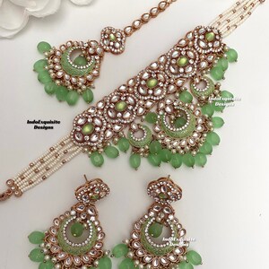 May include: A green and gold Indian bridal necklace set with a matching pair of earrings. The necklace features a large, ornate pendant with green stones and pearls. The earrings are also decorated with green stones and pearls.