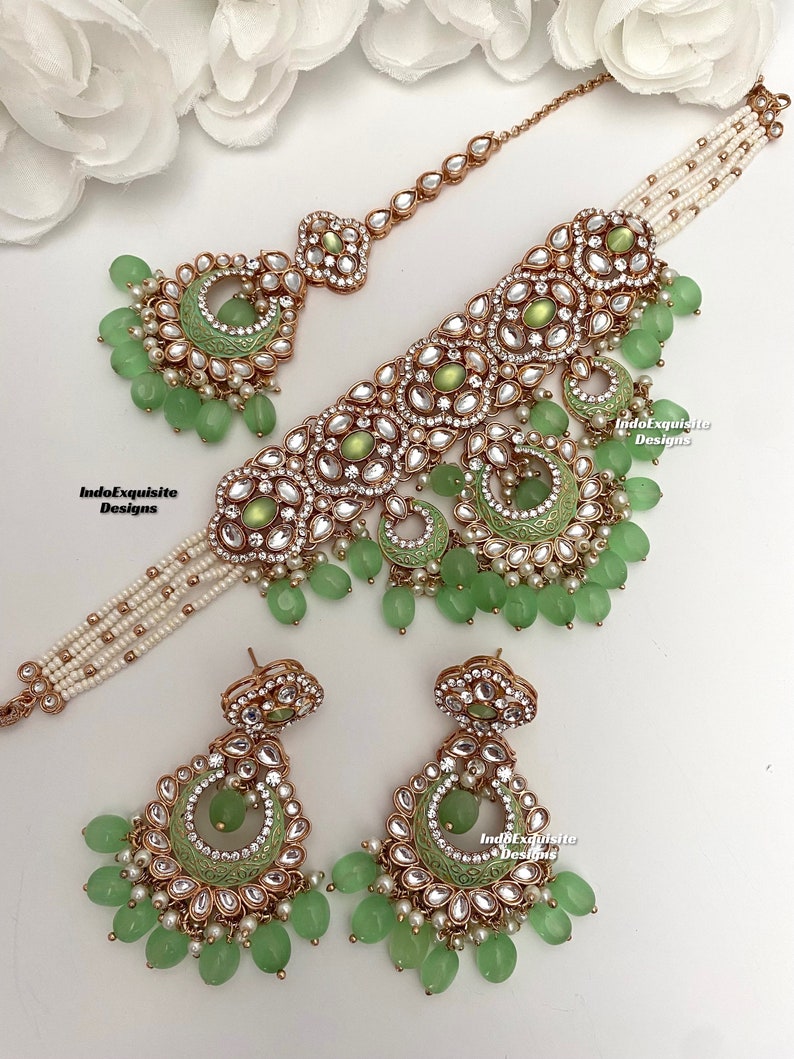 May include: A green and gold Indian bridal necklace set with a matching pair of earrings. The necklace features a large, ornate pendant with green stones and pearls. The earrings are also decorated with green stones and pearls.