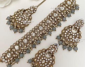 May include: A gold and silver necklace set with a choker style necklace, matching earrings, and a maang tikka. The set features a design of round and oval-shaped stones with a gray bead fringe.
