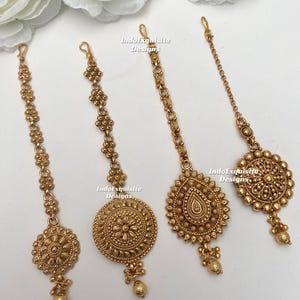 Op de afbeelding: Vier goudkleurige Indiase haarstukken, of maang tikkas, met ingewikkelde bloemen- en geometrische ontwerpen. Elk stuk heeft een centrale hanger en een ketting of draad voor bevestiging. De tekst "IndoExquisite Designs" is zichtbaar.
