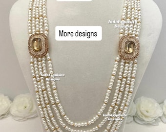 Op de afbeelding: Een parelketting met meerdere strengen, met een grote, vierkante, goudkleurige hanger met een amberkleurige steen. De ketting wordt op een mannequin getoond. Tekst op de afbeelding luidt "Design 1" en "More designs".