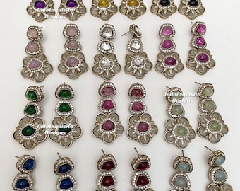 Puede incluir: Una colección de veinticuatro pendientes colgantes dispuestos en cuatro filas, cada uno con tres piedras apiladas de varios colores en metal dorado, detalles de cristales claros y una base floral con el texto IndoExquisite Designs.