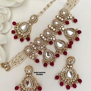 Puede incluir: Un conjunto de collar y pendientes de oro y blanco con cuentas rojas. El collar tiene varias hileras de perlas y un gran colgante con un diseño de espejo. Los pendientes tienen forma de lágrima con un diseño de espejo y cuentas rojas.