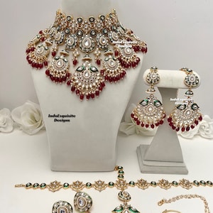 Op de afbeelding: Een traditionele Indiase bruidssieradenset met een gouden ketting, oorbellen, een maang tikka, een neusring en een bijpassende armband. De set is versierd met witte stenen, rode kralen en groene accenten.