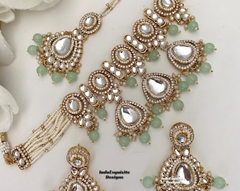 Puede incluir: Un conjunto de collar de oro y plata con un collar estilo choker, pendientes a juego y un maang tikka. El collar presenta múltiples hileras de perlas y un gran colgante con un acabado de espejo. Los pendientes y el maang tikka también presentan detalles de espejo y están adornados con cuentas verdes.