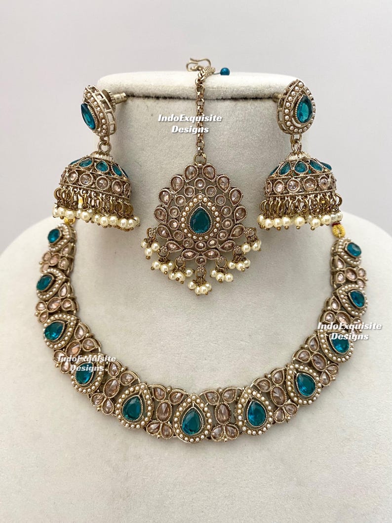 May include: An antique gold-toned jewelry set featuring a necklace, earrings, and a headpiece. The necklace and earrings have teardrop-shaped teal-colored stones. The earrings are bell-shaped with small white pearls. The headpiece has a central teal stone and pearl accents.