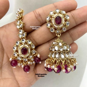 May include: A pair of gold-tone earrings with a dangling design. The earrings feature a round, floral-shaped top with a red gemstone in the center. The bottom of the earrings have a cluster of pink and white beads.