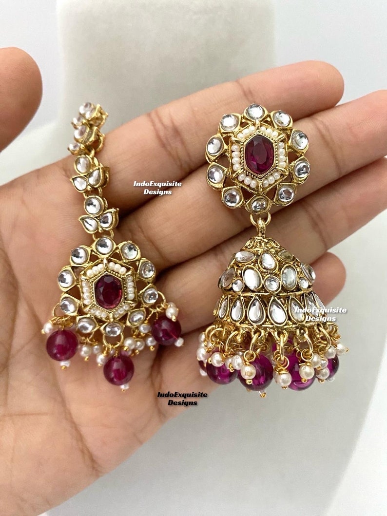 May include: A pair of gold-tone earrings with a dangling design. The earrings feature a round, floral-shaped top with a red gemstone in the center. The bottom of the earrings have a cluster of pink and white beads.