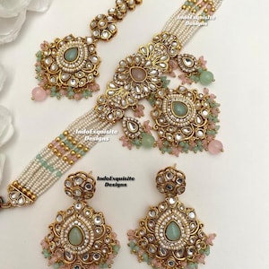 Puede incluir: Un conjunto de joyería dorado que incluye una gargantilla de perlas de varias hebras con cuentas verde menta y rosas, un maang tikka a juego y dos pendientes colgantes. Cada pieza está adornada con piedras claras y acentos en forma de gota verde menta. Texto: IndoExquisite Designs.