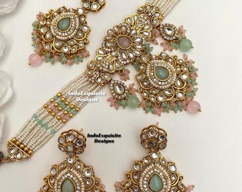 May include: A gold-toned jewelry set featuring a multi-strand pearl choker with mint green and pink beads, a matching maang tikka, and two drop earrings. Each piece is adorned with clear stones and central mint green teardrop accents. Text reads IndoExquisite Designs.