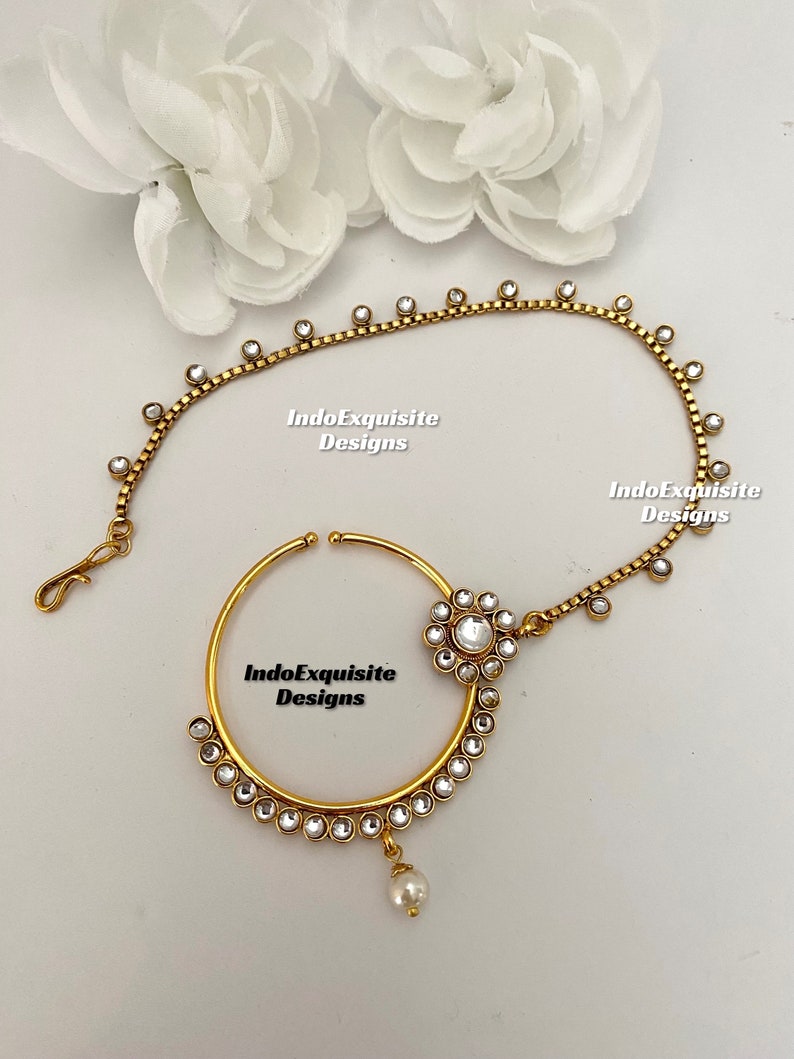 May include: Gold nose ring with a pearl drop and a chain with rhinestones. The chain is attached to the nose ring and has a flower-shaped design with rhinestones.