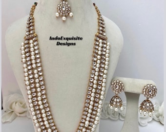 May include: A four-strand gold necklace with white pearls and clear stones. The necklace is paired with matching earrings and a matching tikka.