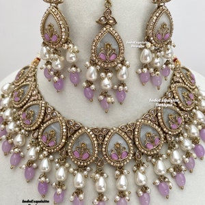 May include: A detailed jewellery set comprising a necklace, earrings, and a headpiece. The gold-toned metal set features teardrop pendants with white centers, pink floral details, white faux pearls, and lavender beads. A beautiful set for special occasions.