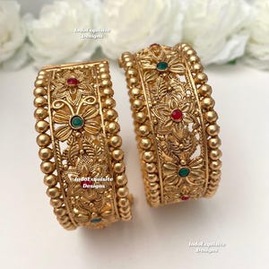 May include: Two gold-colored bangles with intricate floral and leaf designs. Each bangle features red and green gemstone accents. The bangles are adorned with small, round beads along the edges. The text "IndoExquisite Designs" is visible.