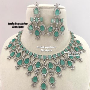 May include: A silver necklace and earrings set with a teardrop design. The necklace features a choker style with a row of emerald green stones and a cascading design of teardrop stones. The earrings are dangle style with a teardrop stone and a row of emerald green stones.