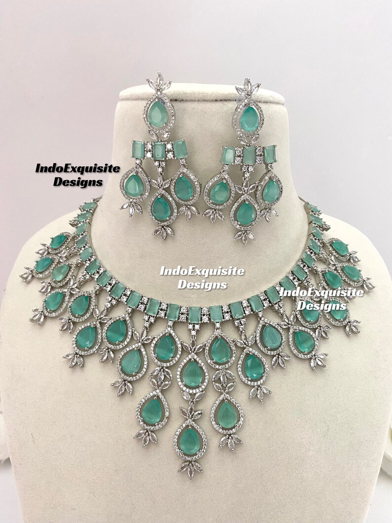 May include: A silver necklace and earrings set with a teardrop design. The necklace features a choker style with a row of emerald green stones and a cascading design of teardrop stones. The earrings are dangle style with a teardrop stone and a row of emerald green stones.