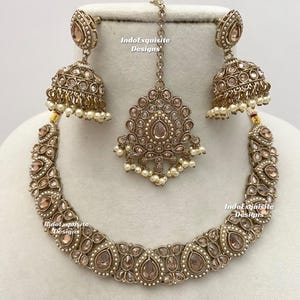 May include: A complete antique gold-toned jewelry set featuring a necklace, earrings, and a headpiece. The set is adorned with clear and peach-colored stones and small white pearls. The text "IndoExquisite Designs" is visible on the image.