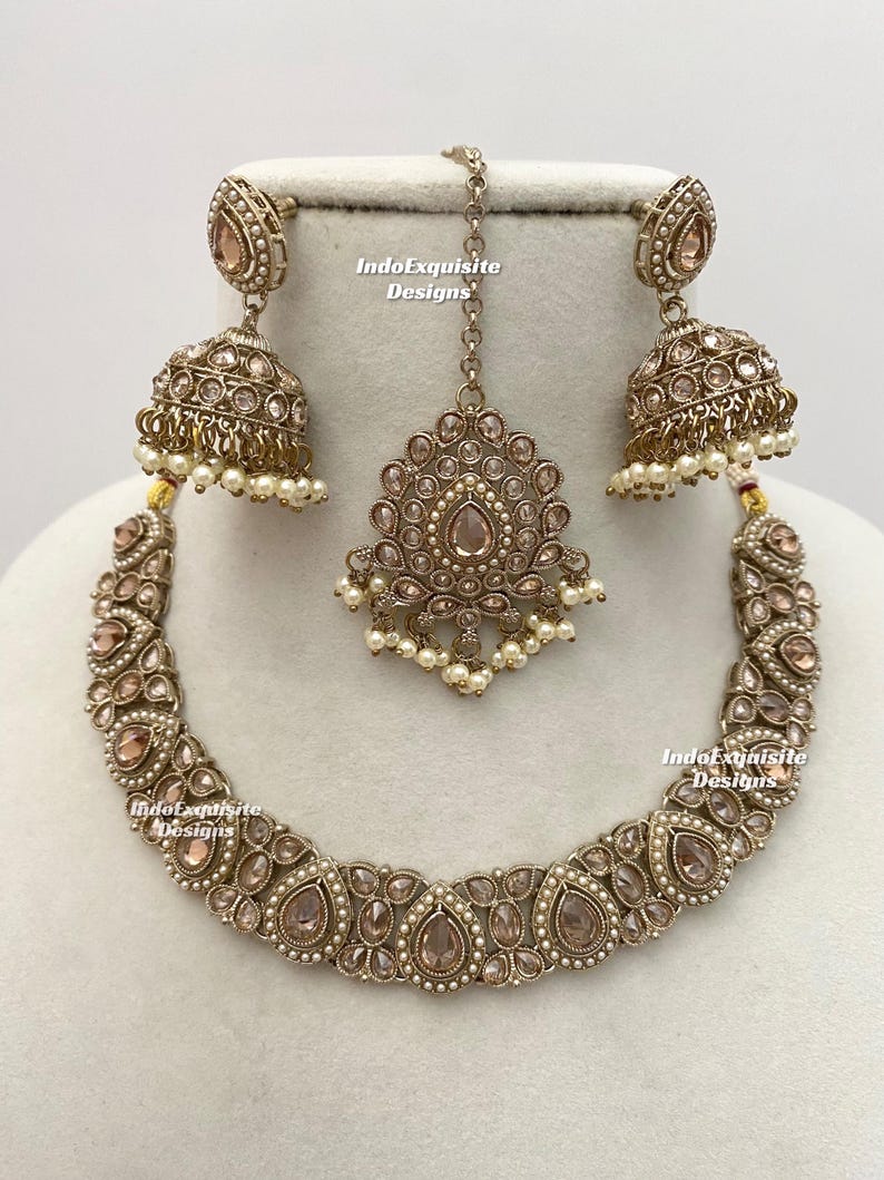 May include: A complete antique gold-toned jewelry set featuring a necklace, earrings, and a headpiece. The set is adorned with clear and peach-colored stones and small white pearls. The text "IndoExquisite Designs" is visible on the image.