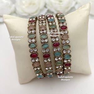 Pode incluir: Quatro pulseiras douradas com pedras preciosas ovais coloridas em vermelho, rosa, verde-azulado e transparente. As pulseiras são exibidas em uma almofada branca com o texto "IndoExquisite Designs". As pedras são dispostas em um padrão repetitivo.