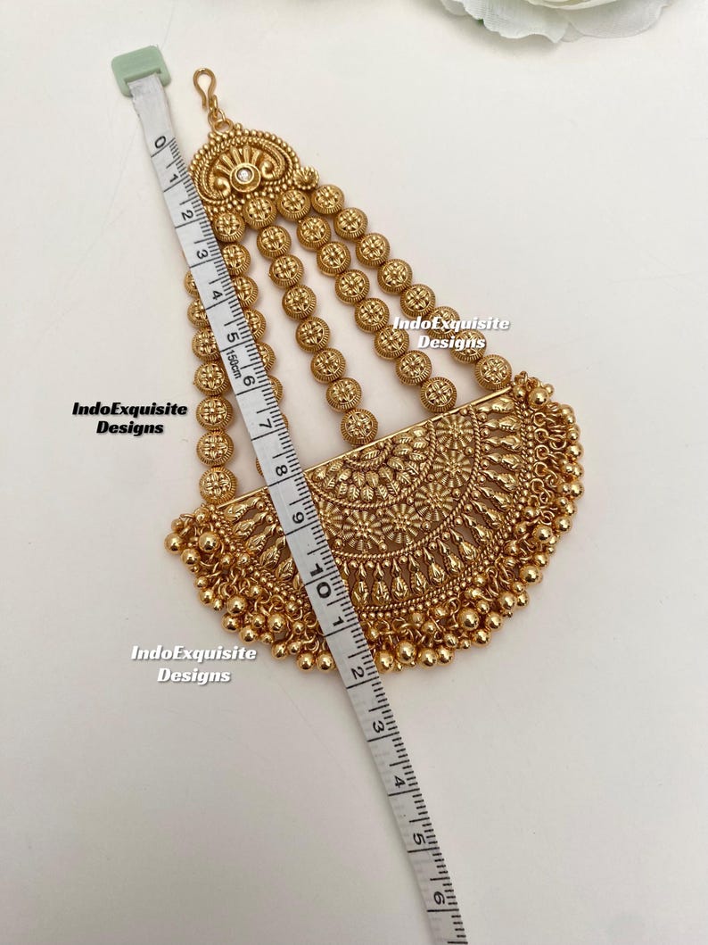 May include: A gold-colored headpiece with intricate floral and beaded designs. The headpiece is measured with a tape measure, showing a length of approximately 5 inches. The text "IndoExquisite Designs" is visible.
