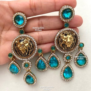 May include: A pair of gold tone earrings with a lion head design and turquoise blue crystal accents. The earrings feature a large round pendant with a lion head design, surrounded by smaller turquoise blue crystals. The lion head is gold tone and has a black enamel background. The earrings are suspended from a smaller round pendant with a turquoise blue crystal.